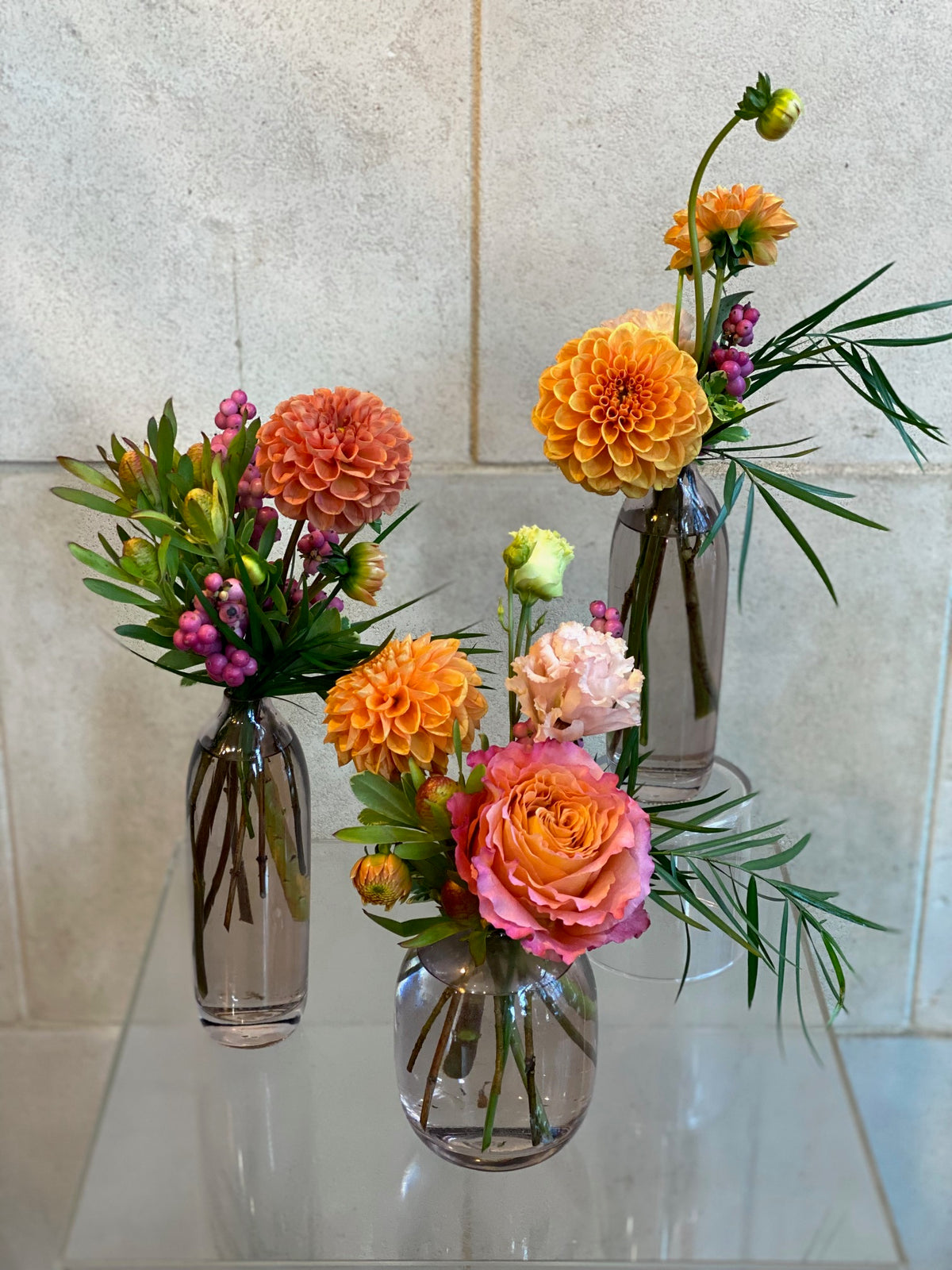 Set of three glass bud vases with seasonal flowers and greenery in warm autumn tones.