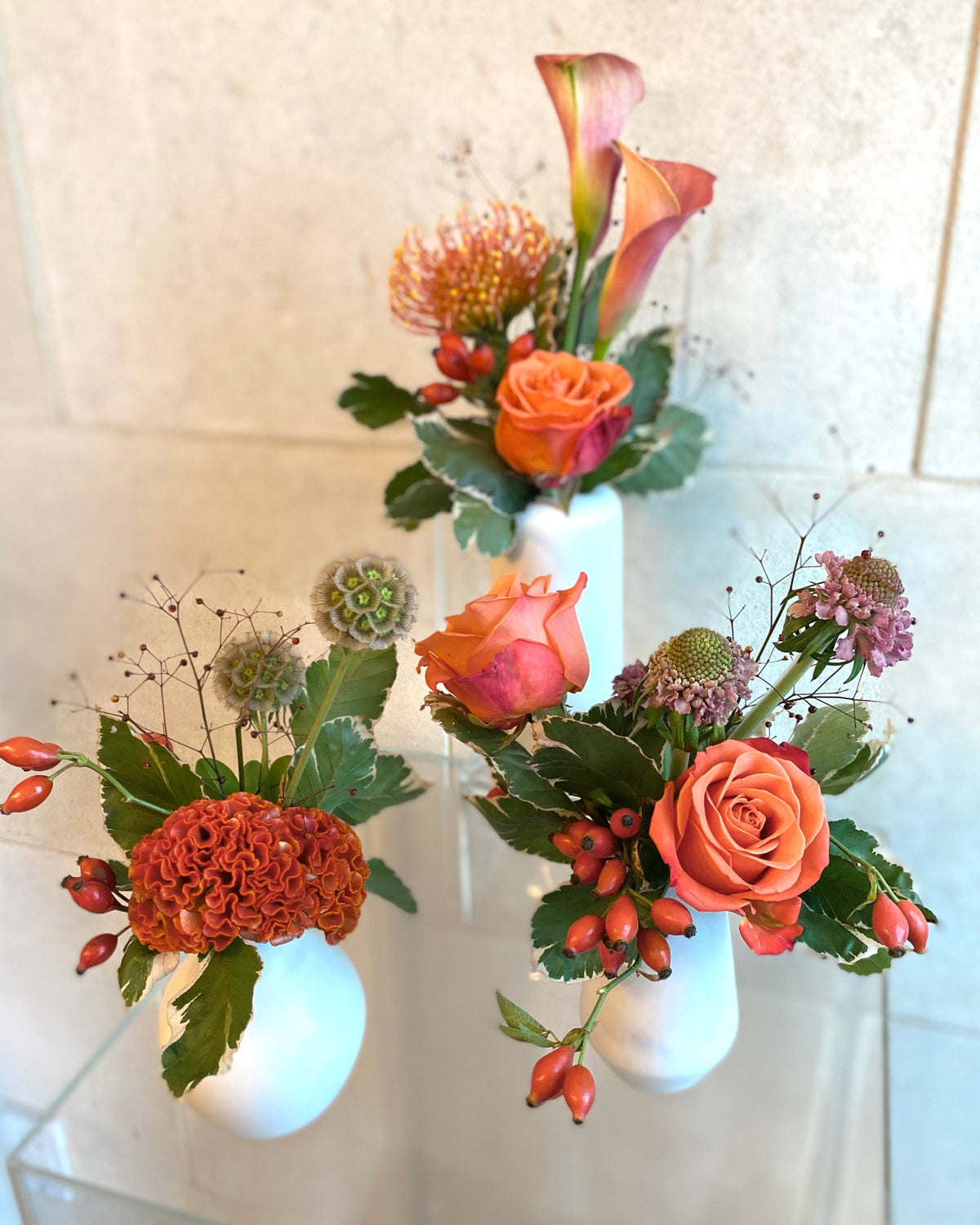 Set of three white ceramic bud vases with seasonal flowers in warm orange and coral tones